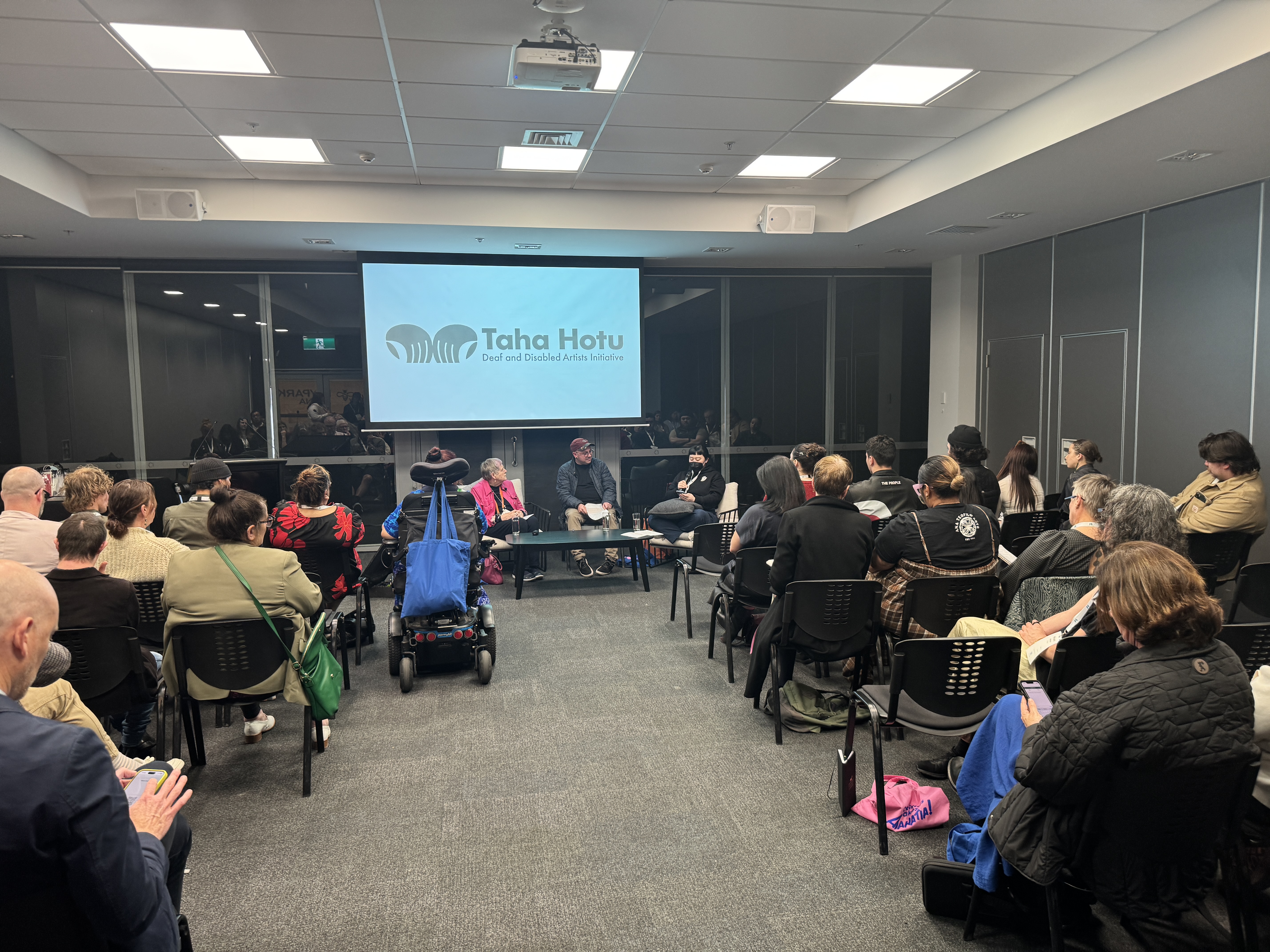 A large crowd sits in a gathering before a projector screen which features the Taha Hotu logo. Robyn, Stace and Kim sit facing the crowd, presenting.